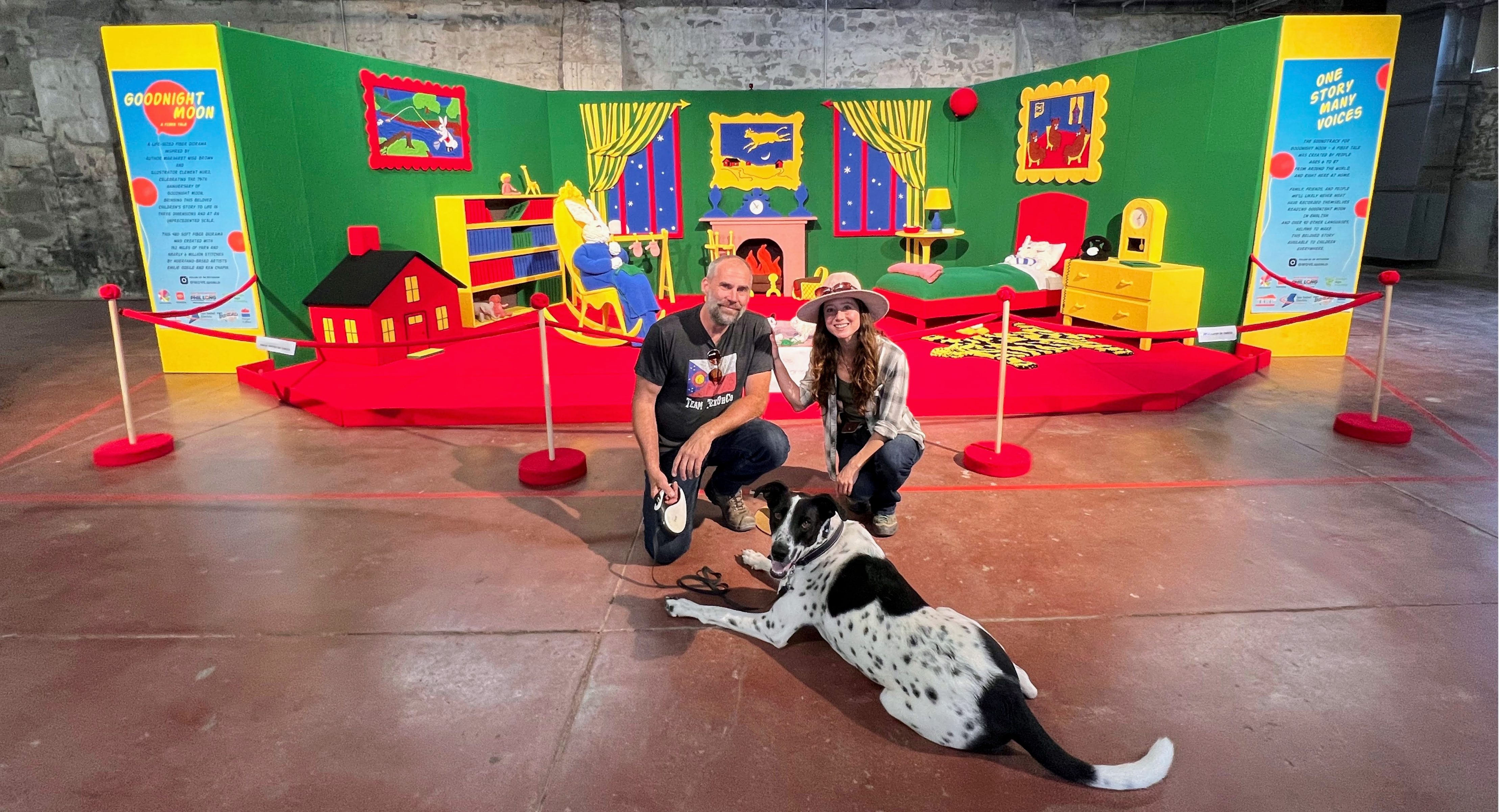 Two people and a dog in front of a Goodnight Moon scene