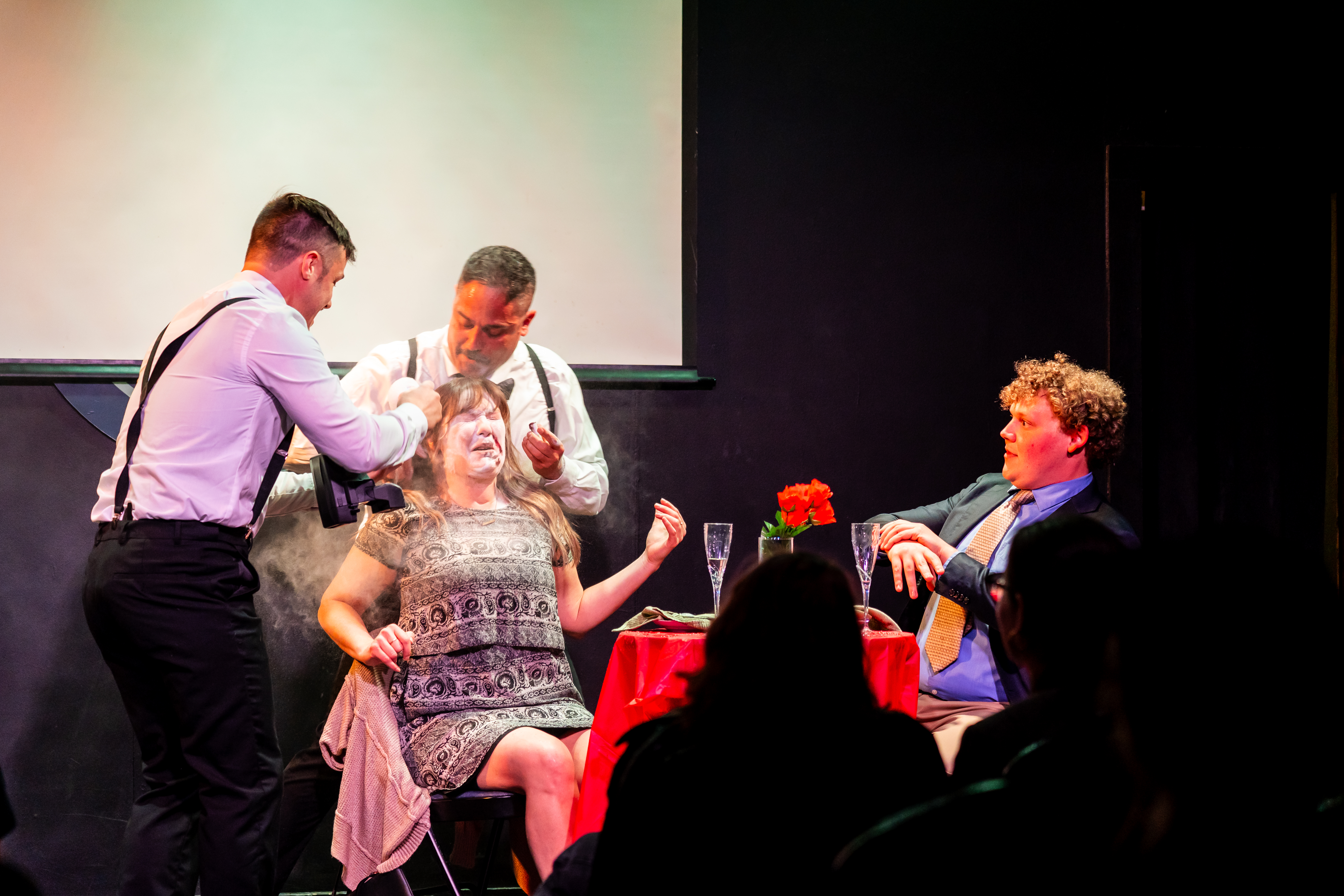 A woman is uncomfortable as two men in suits surrond her and a man in a tie looks confused across the table.