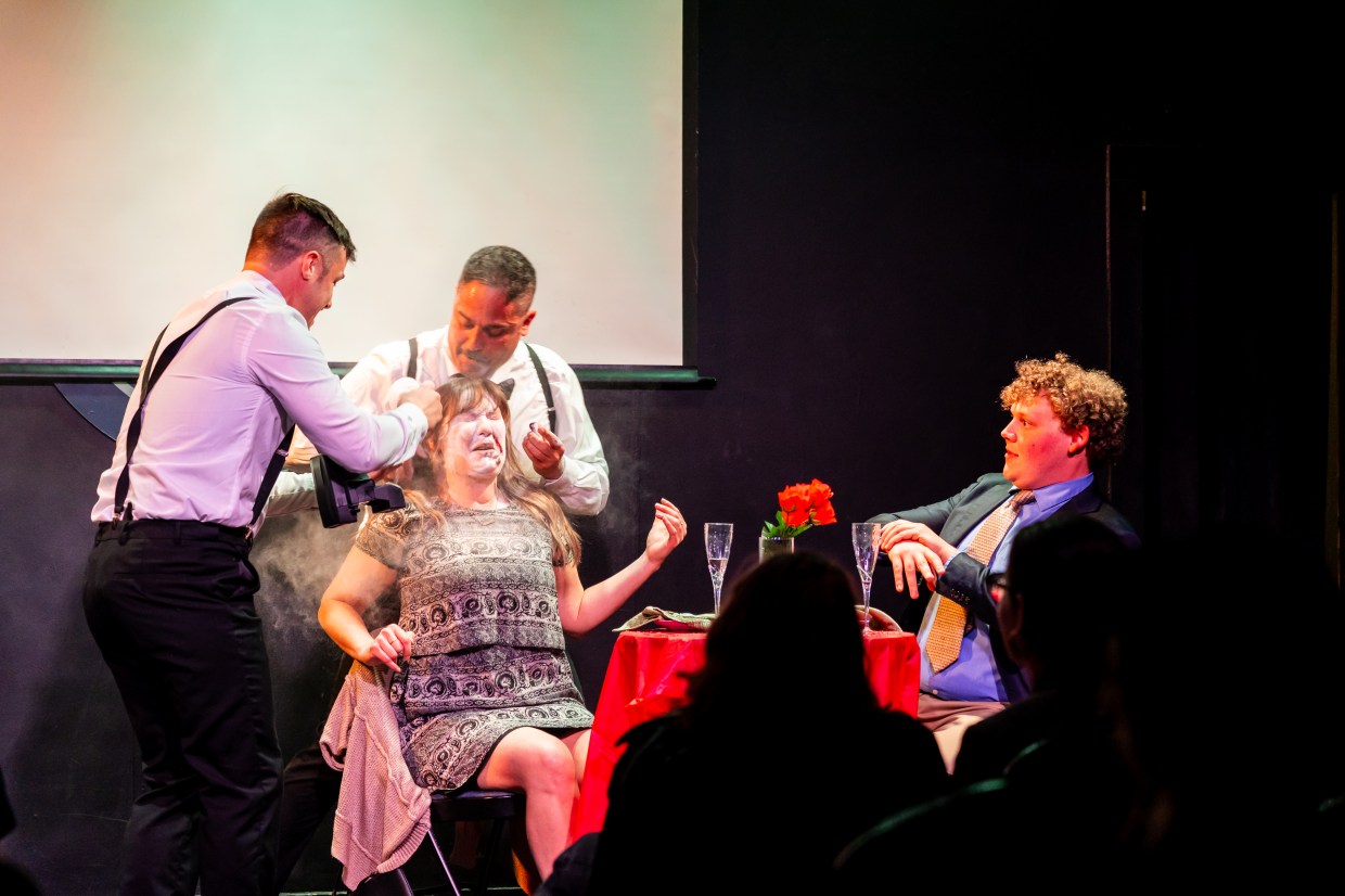 A woman is uncomfortable as two men in suits surrond her and a man in a tie looks confused across the table.