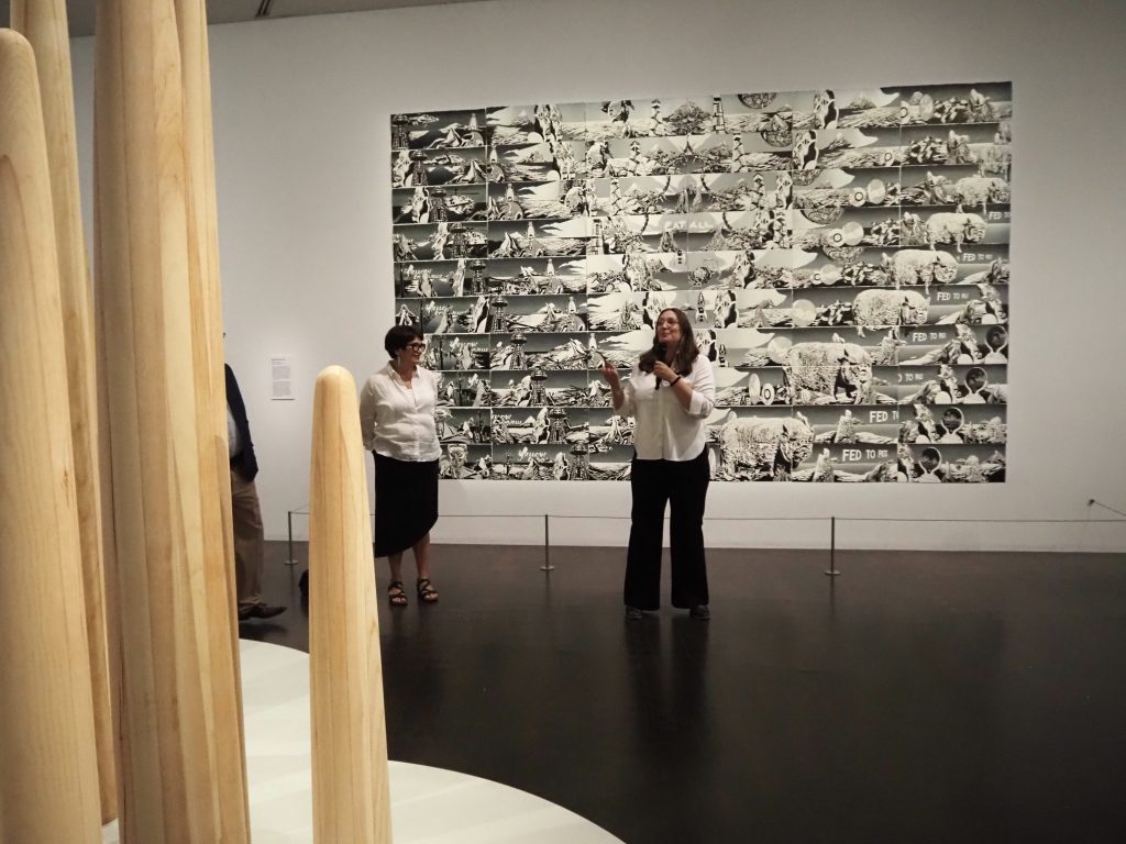A woman speaks, standing before a large black and white artwork.