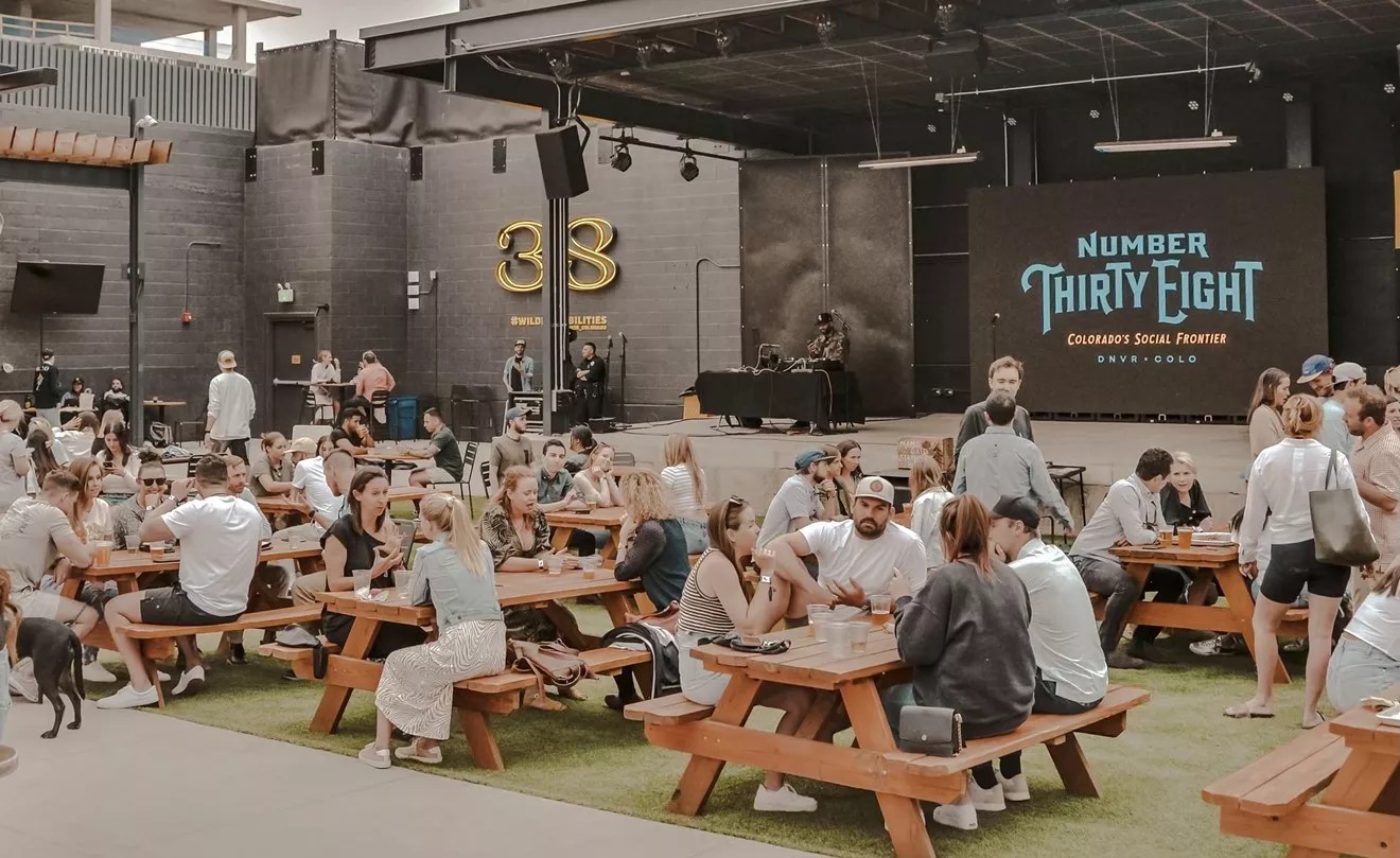 A mix of people in their mid-thirties sits at picnic tables in front of an outdoor stage displaying a projected banner with "Number Thirty Eight" in large letters.