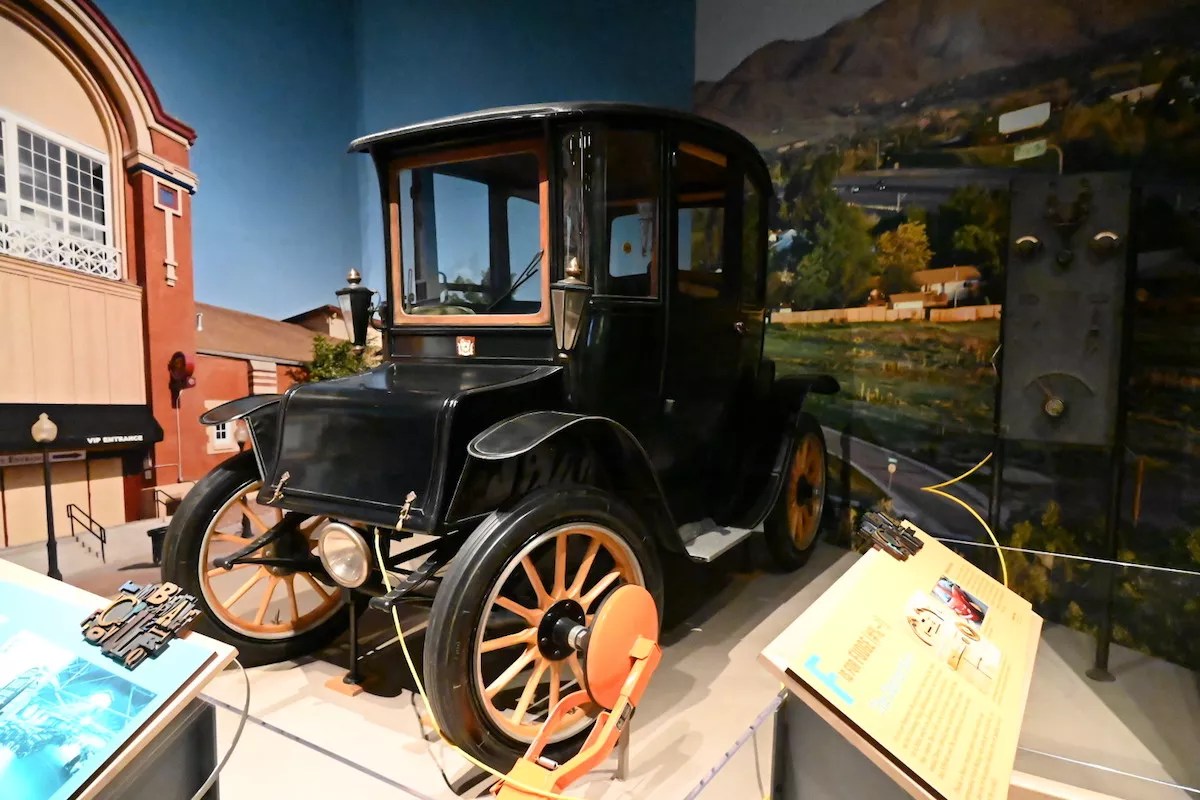 A 1914 Fritchle Electric Colonial Coupe model 283 in History Colorado in front of its charging station with another Denver invention, the boot.