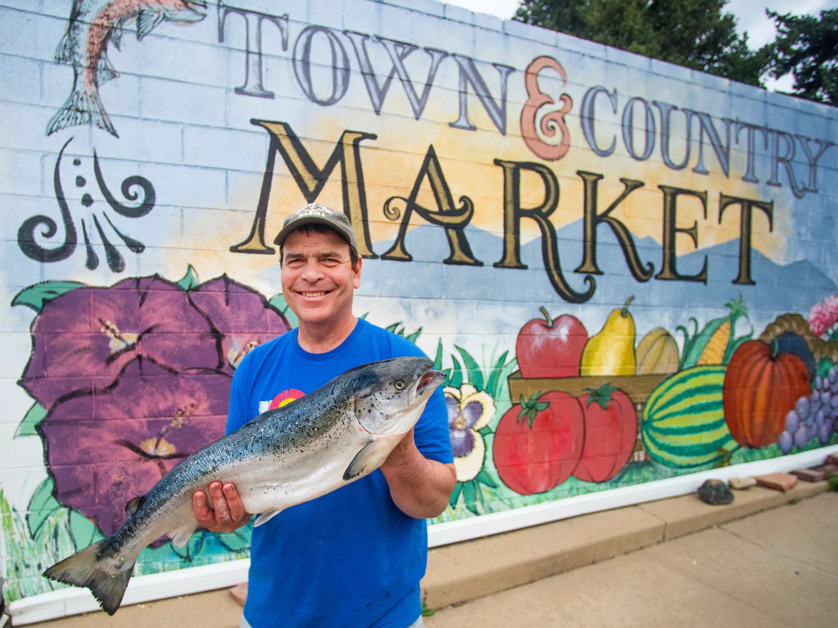 a man holds a large salmon in front of a colorful mural with fruits and vegetables
