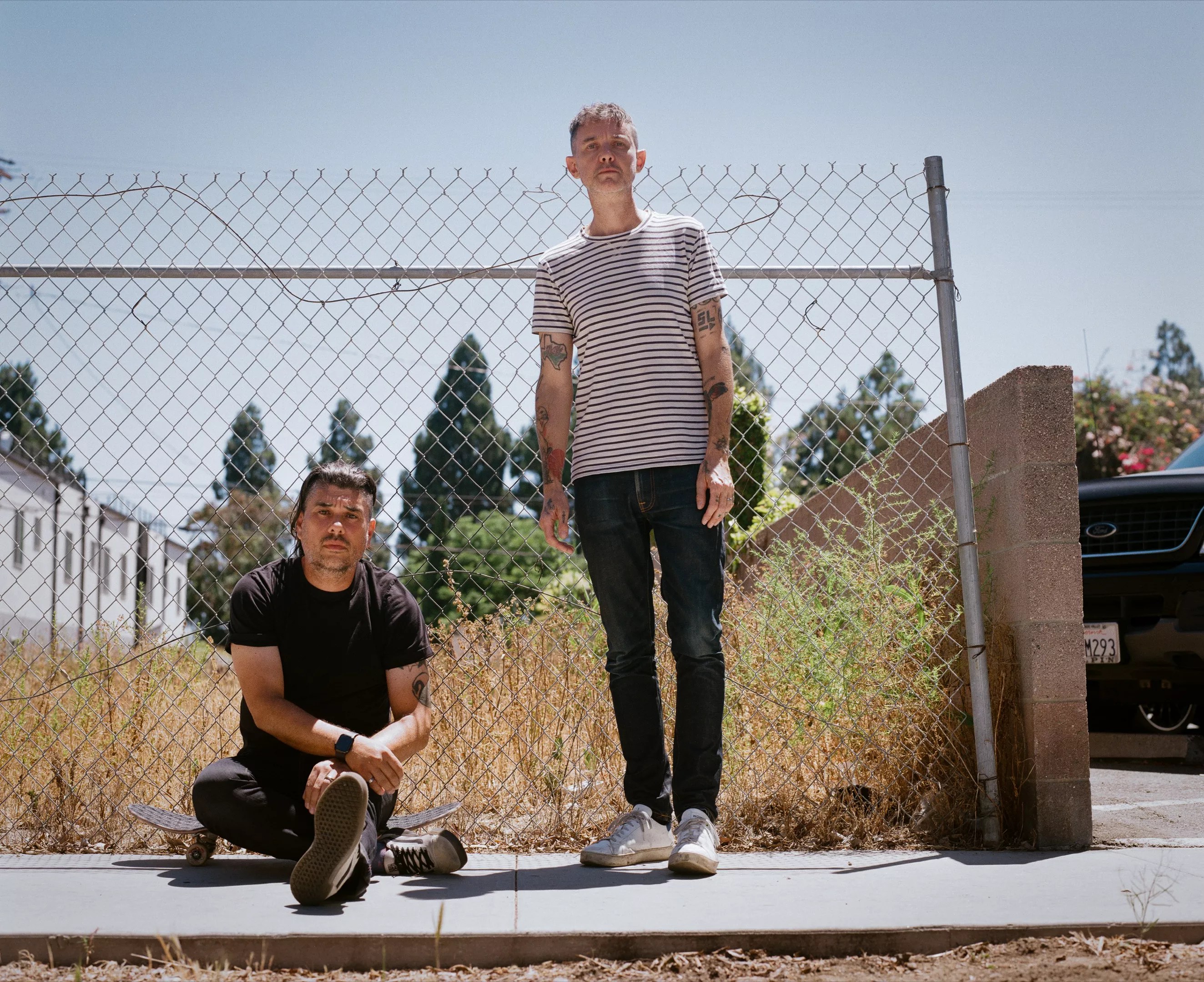 two men pose in front of a chainlink fence