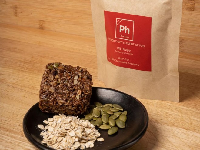 a brown package next to a plate with a trail bar and a pile of seeds and oats