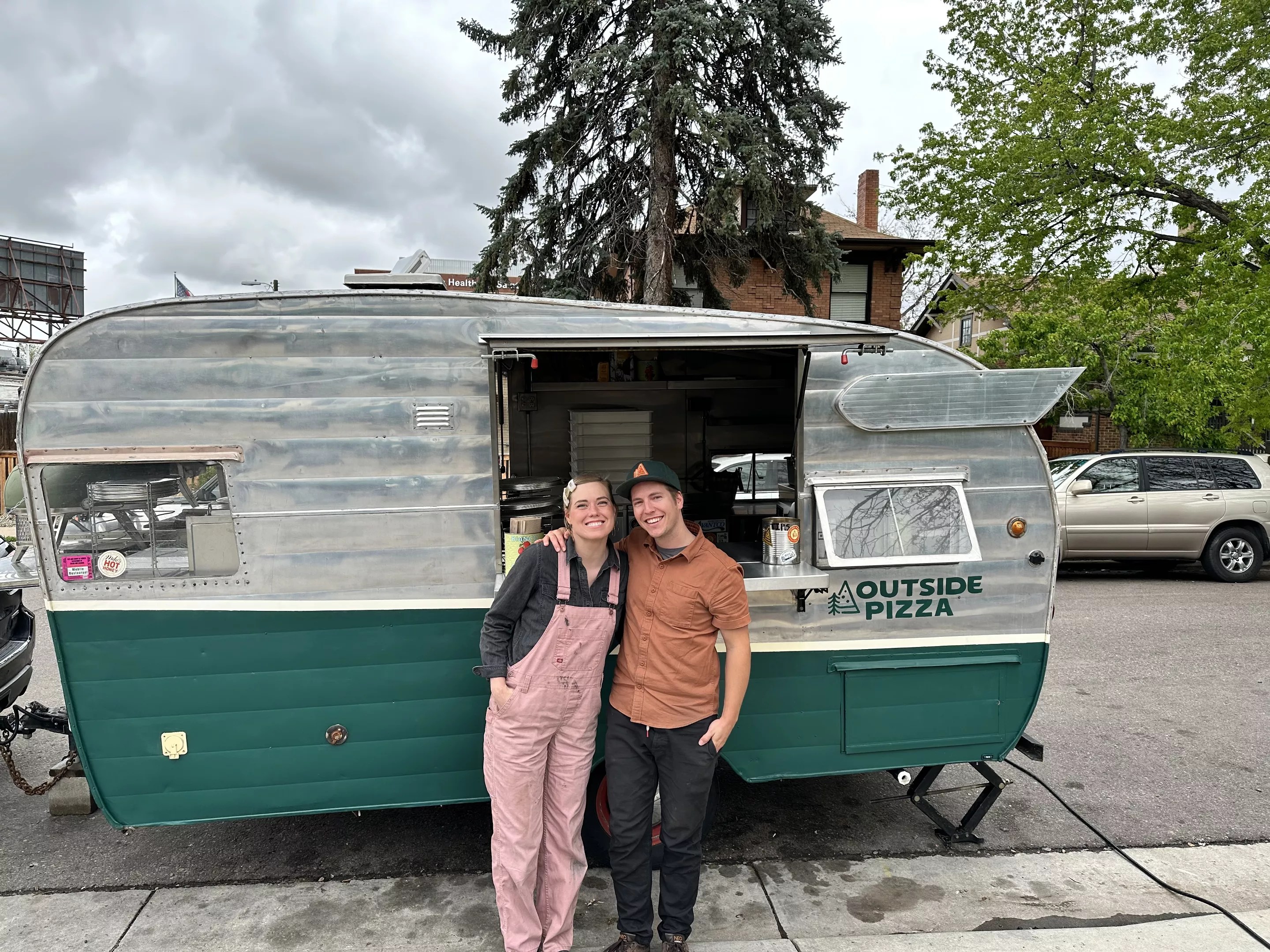 a woman in pink overalls and a man in a brown shirt stand in front of a retro green and white camper