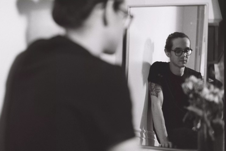 man with glasses and black t-shirt looks in mirror.