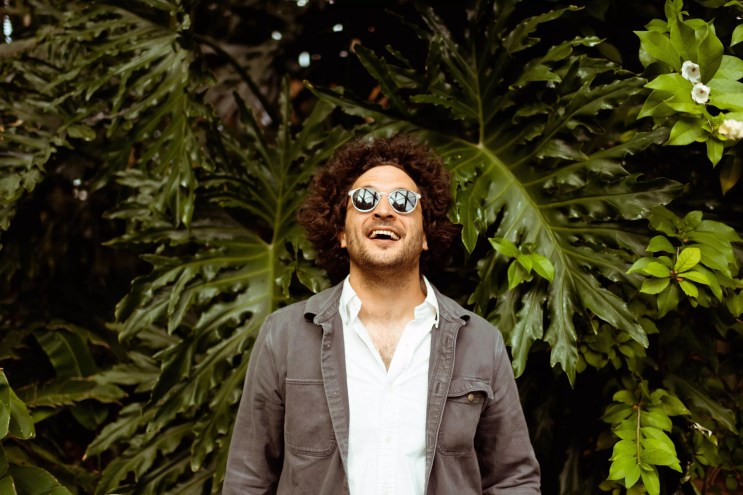 man in sunglasses smiles in front of evergreen tree.