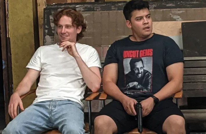 a ginger boy in a white t shirt and a man with black hair in a black shirt holding a fake gun.