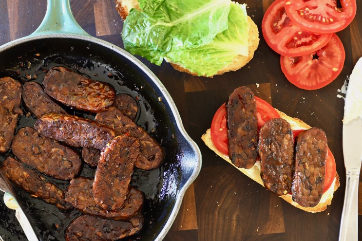 a pan with slices of tempeh next to lettuce and tomatoes