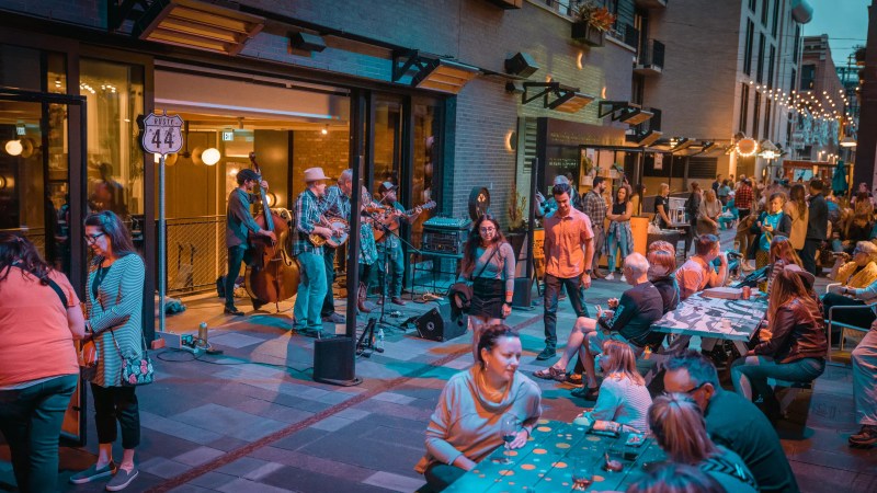 Many people seated with food and drink or walking down a dusk-lit alley while an acoustic band plays on the left.
