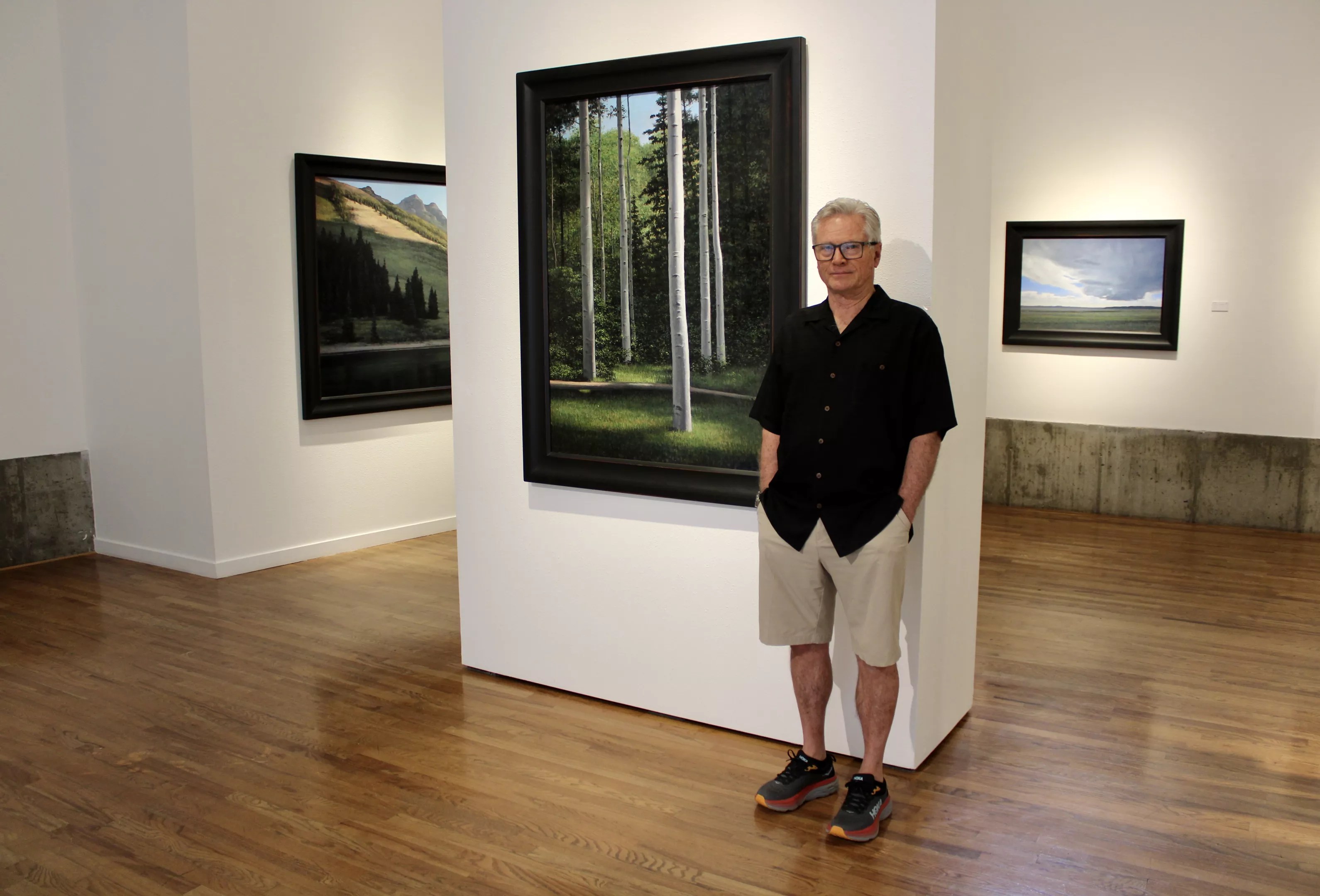 man in black shirt and khaki shorts poses in a gallery with landscape paintings.