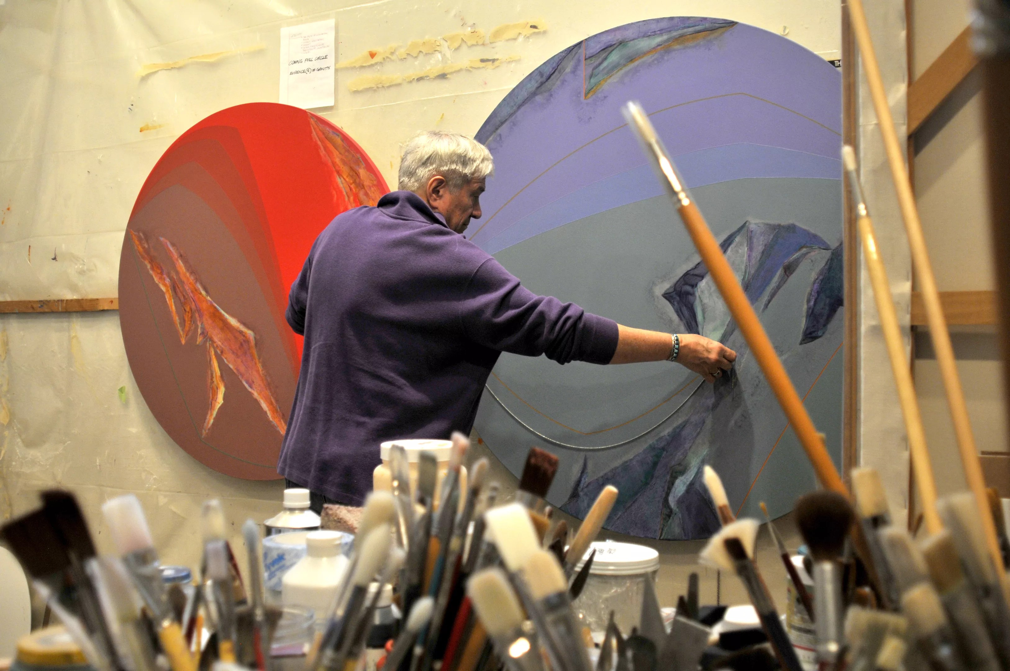 Older man painting on large circular canvas in art studio.