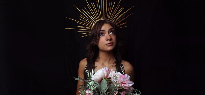 brunette woman wearing gold crown and holding pink flowers.