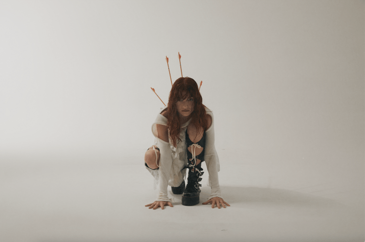 woman crouches in white dress with arrows sticking out of her back