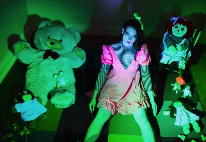 woman dressed as a doll in green lighting surrounded by teddy bears.