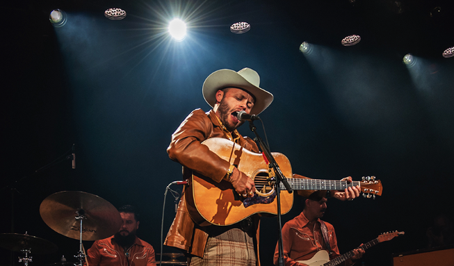 man in cowboy hat singing into mic while playing acoustic guitar.
