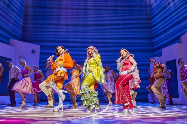 women in orange, yellow and red costumes dance on stage with blue background