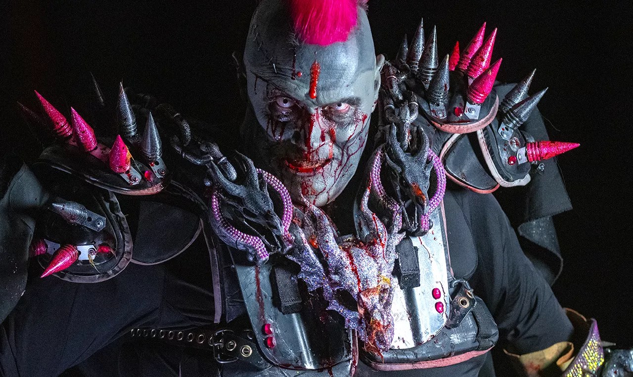 man in zombie facepaint with fake blood has a pink mohawk and is wearing spiky armor