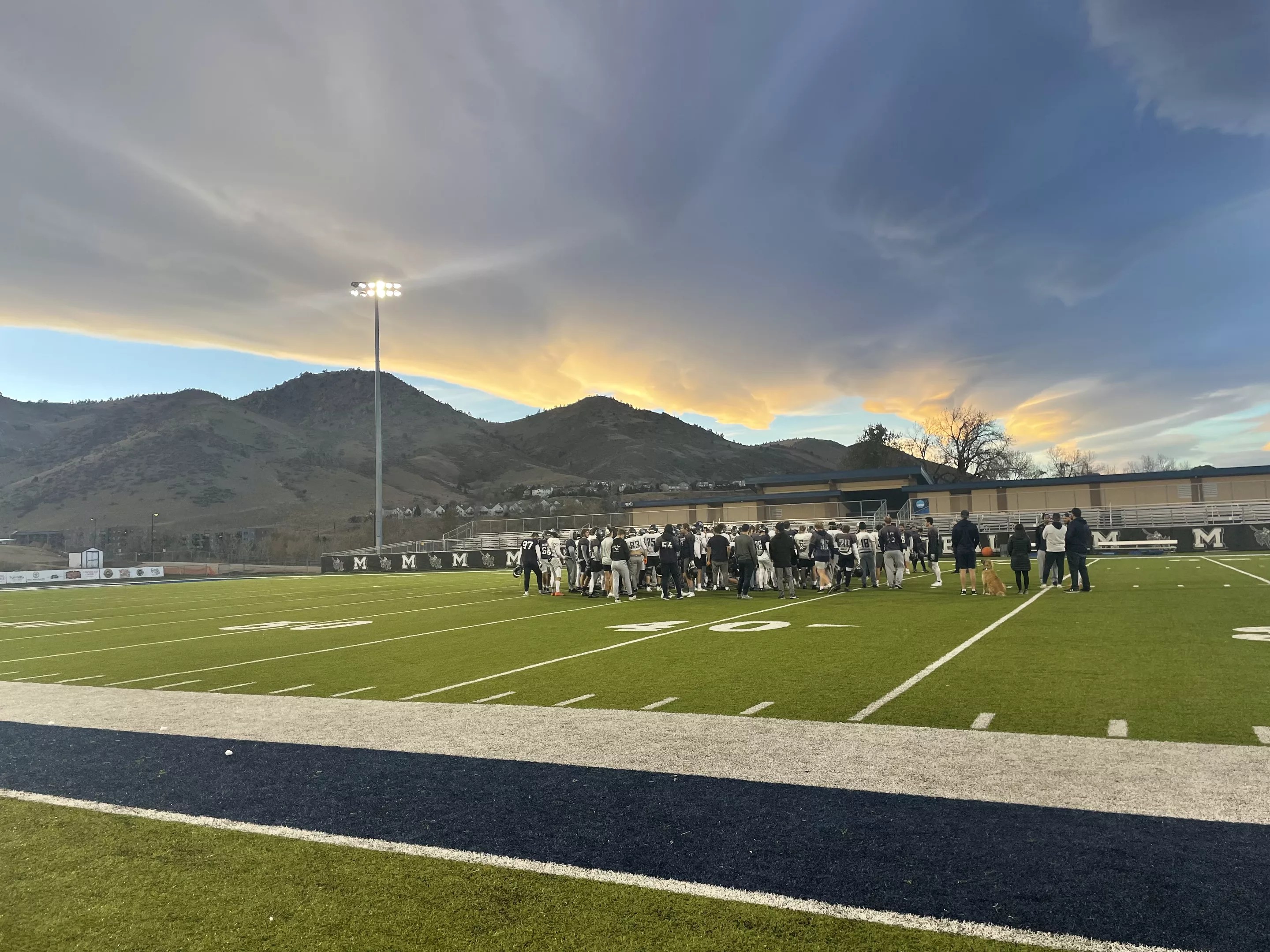 Under a cloudy sky, a football team huddles on a green turf field.