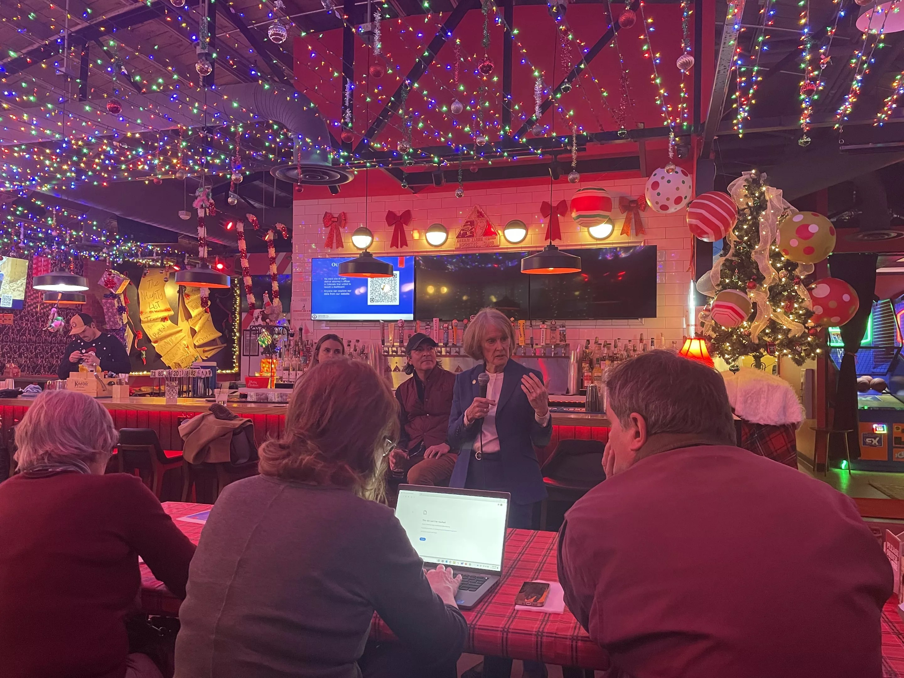 A woman in a blue suit speaks to people gathered in a room decorated for Christmas.