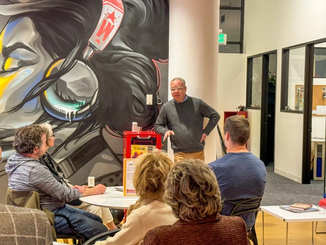 man talking in room with painted mural, people listening.