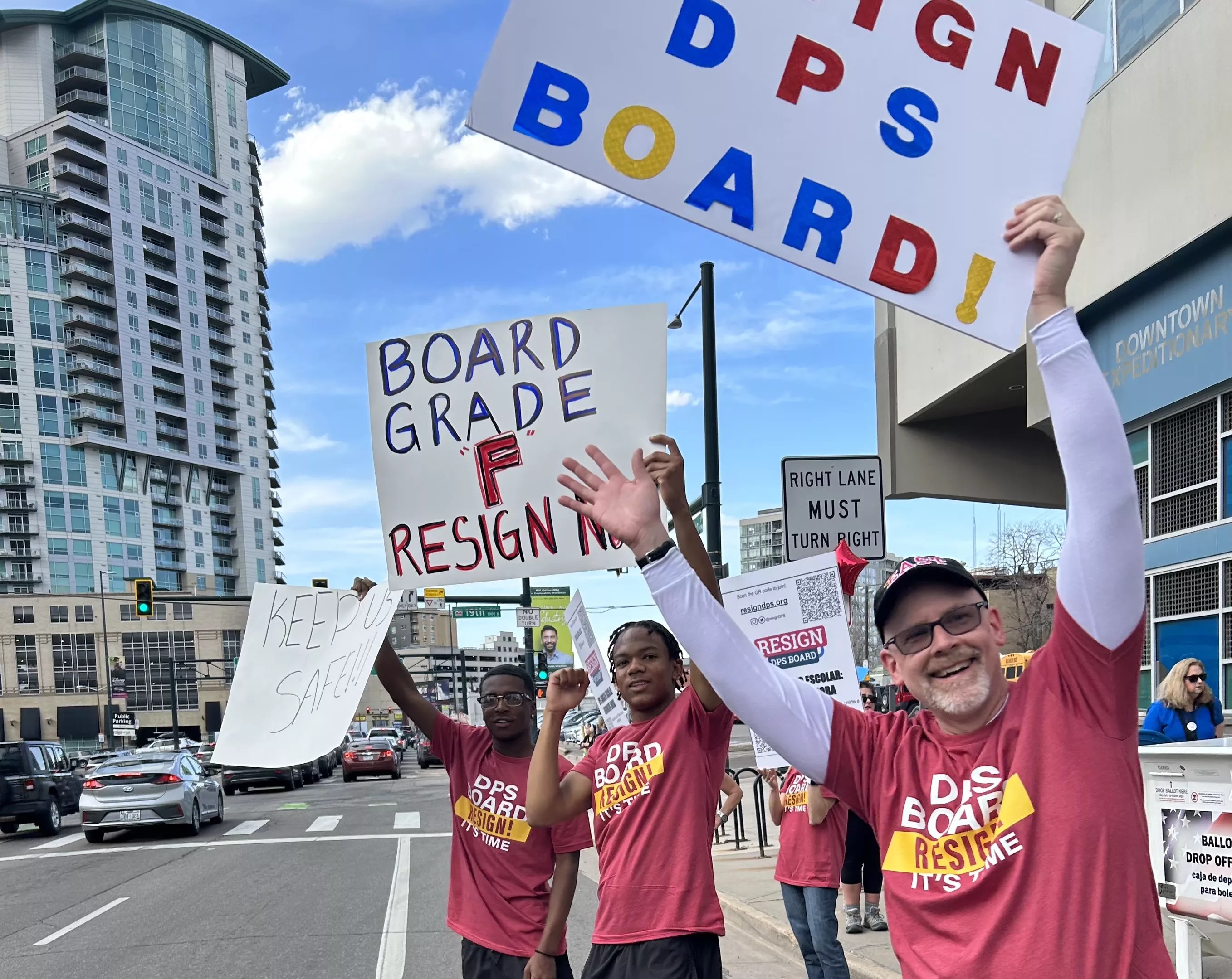 Two students and one parent stand on the side of the road wearing "Resign DPS" shirts and holding signs calling for board members to step down.