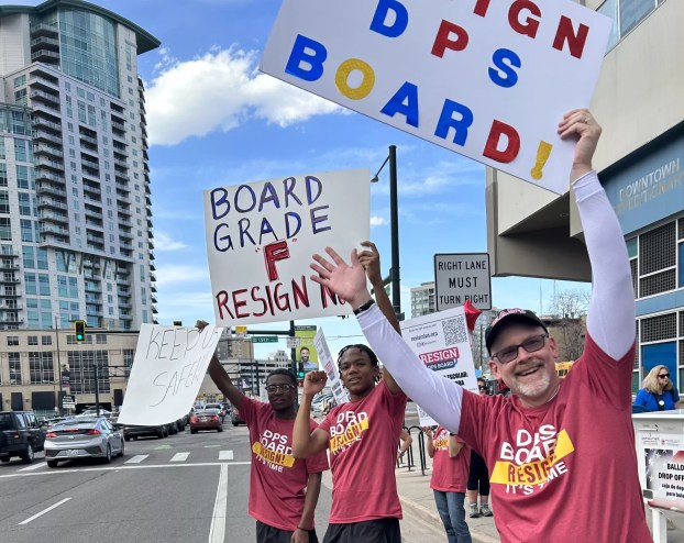 Two students and one parent stand on the side of the road wearing "Resign DPS" shirts and holding signs calling for board members to step down.