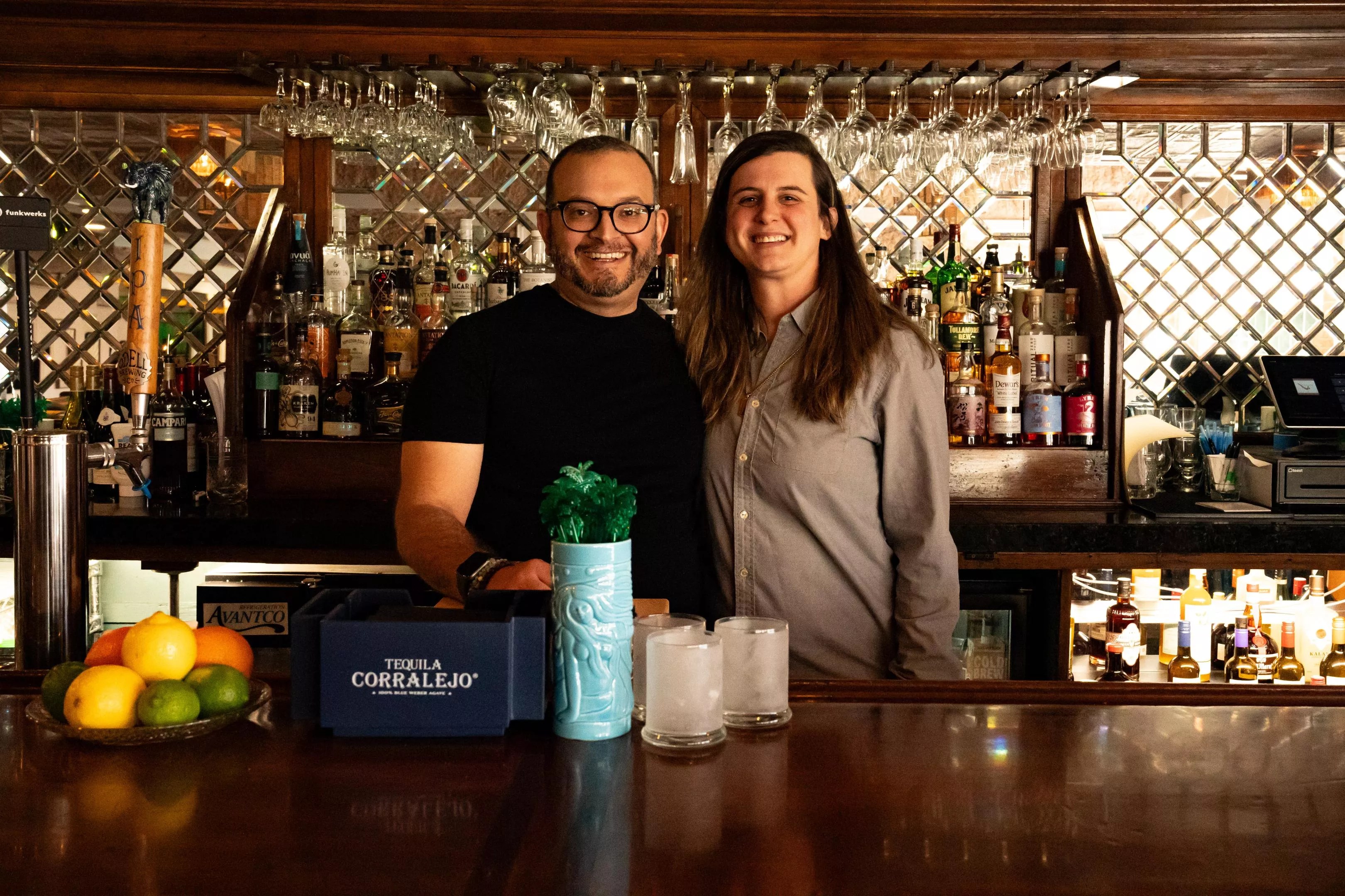 A man in glasses and a black shirt next to a woman in a grey button-down shirt standing behind a bar