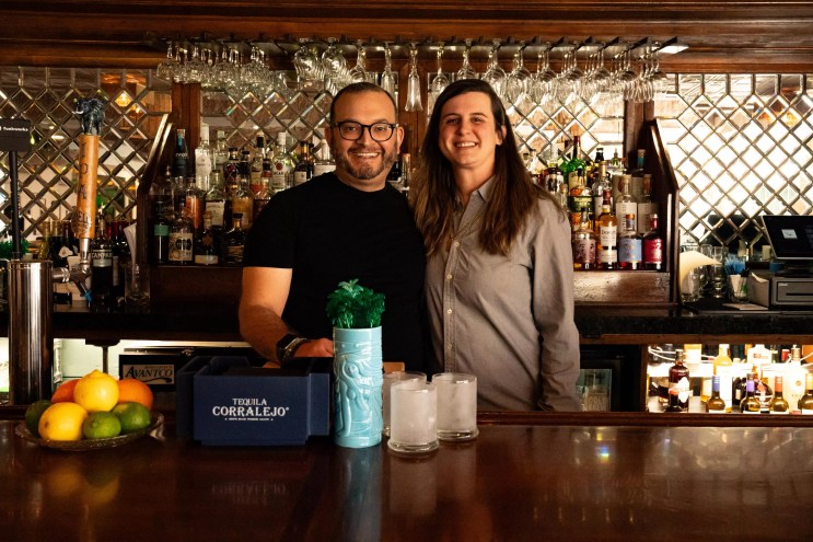 A man in glasses and a black shirt next to a woman in a grey button-down shirt standing behind a bar