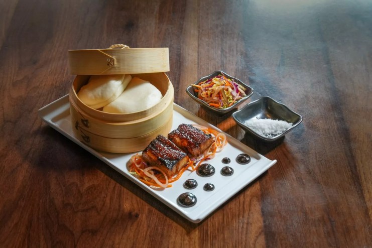 pork belly on a plate next to a steamer basket with bao buns