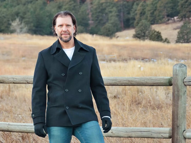 man leans on a fence outdoors wearing jeans and a coat