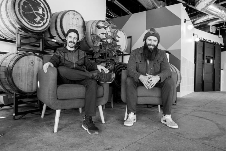 Two men sitting in front of beer barrels.