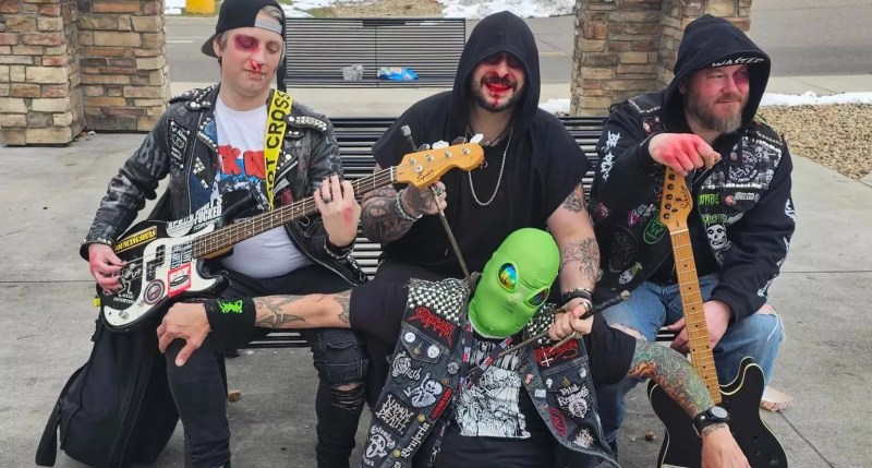 punk bandmates sit on a bench with someone in an alien mask