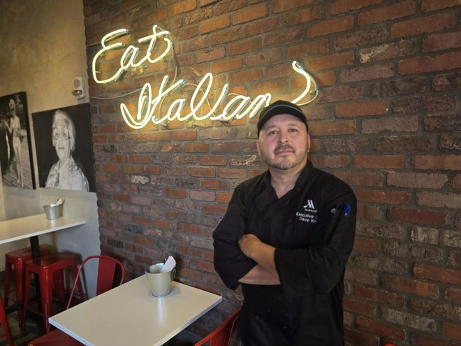 man in a black chef's coat posting in front of a neon "eat Italian" sign