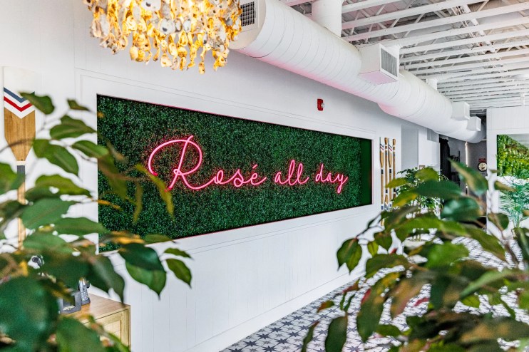 a pink "Rosé All Day" neon sign on a plant-covered background