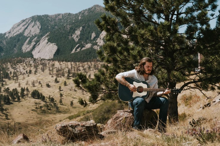 Danno Simpson sits on a rock playing guitar in the Boulder foothills with the Flatirons behind him.
