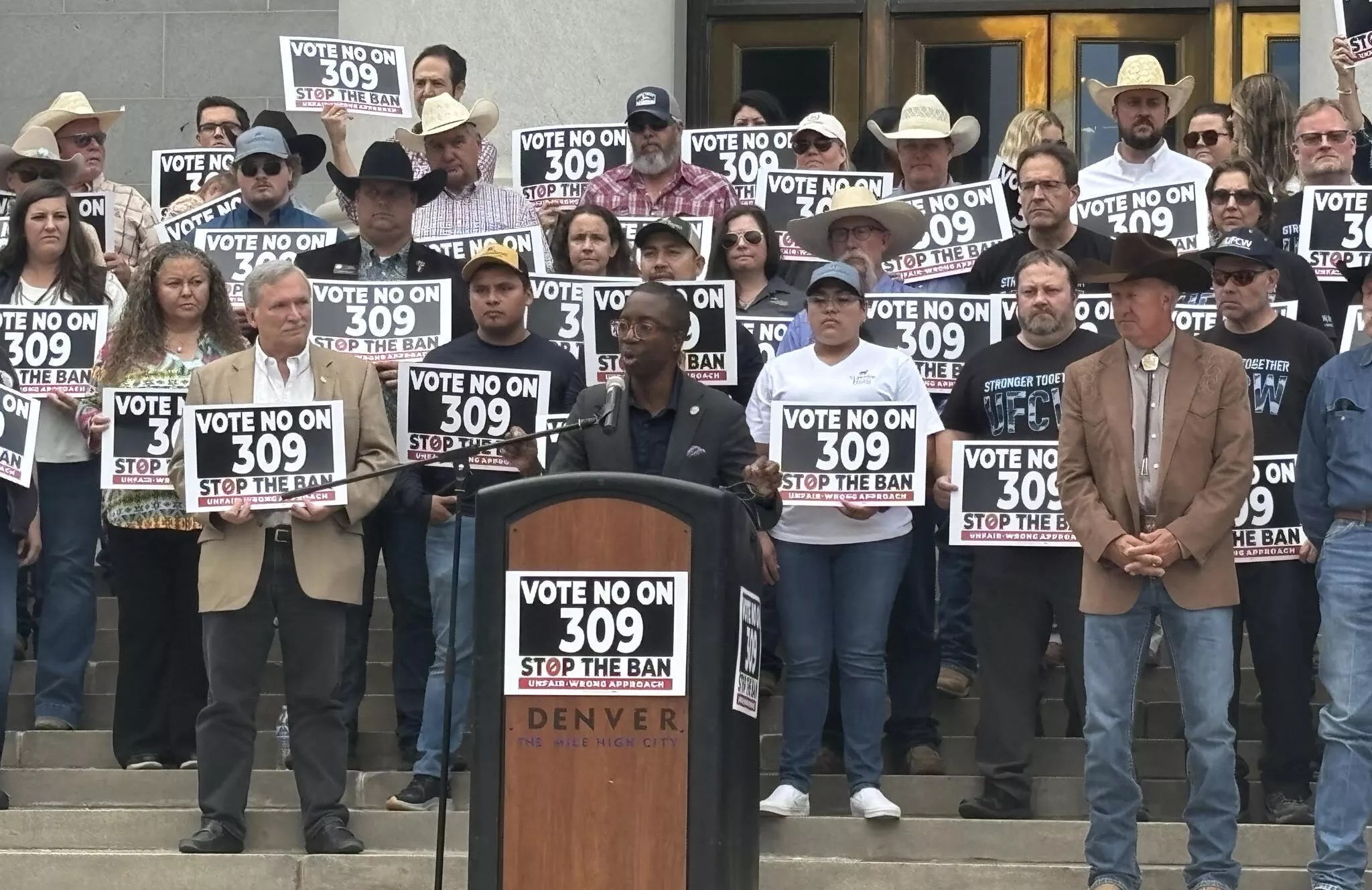 man speaking at podium with crowd behind.