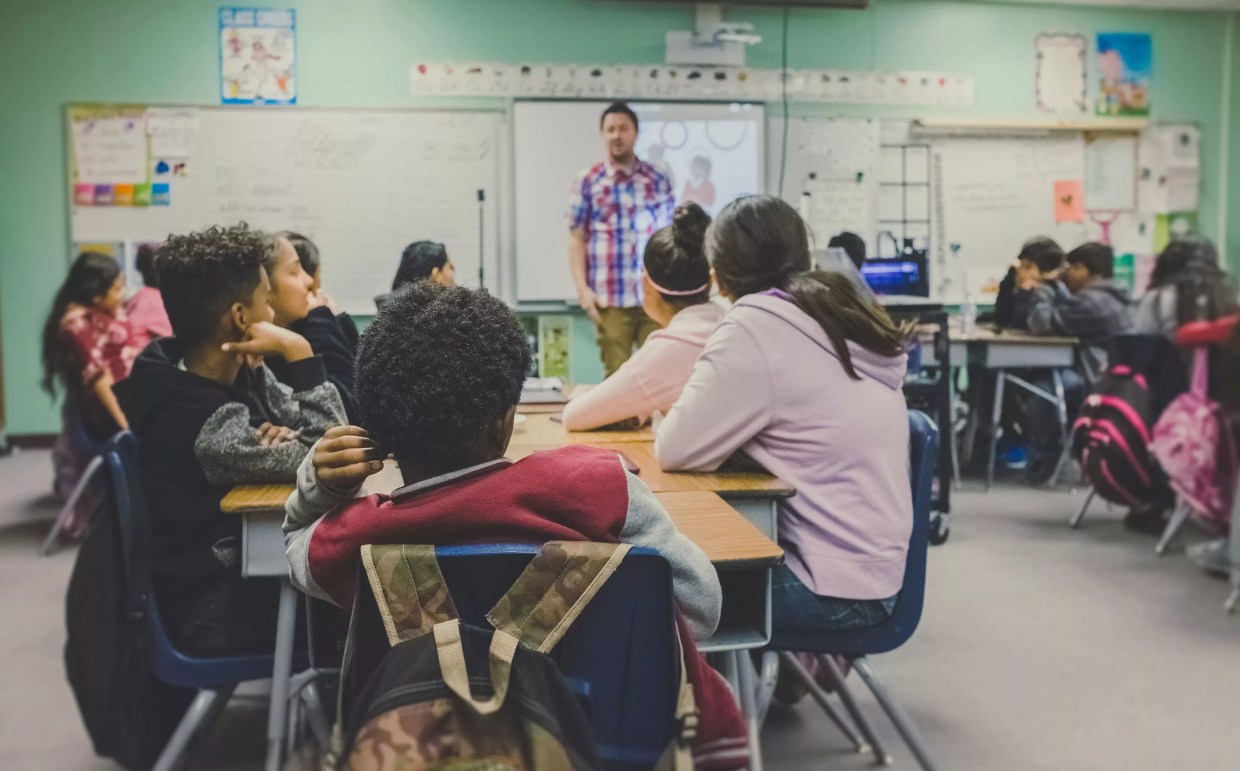 Children sit at desks in a classroom, looking at an adult male teacher standing in front of a whiteboard.