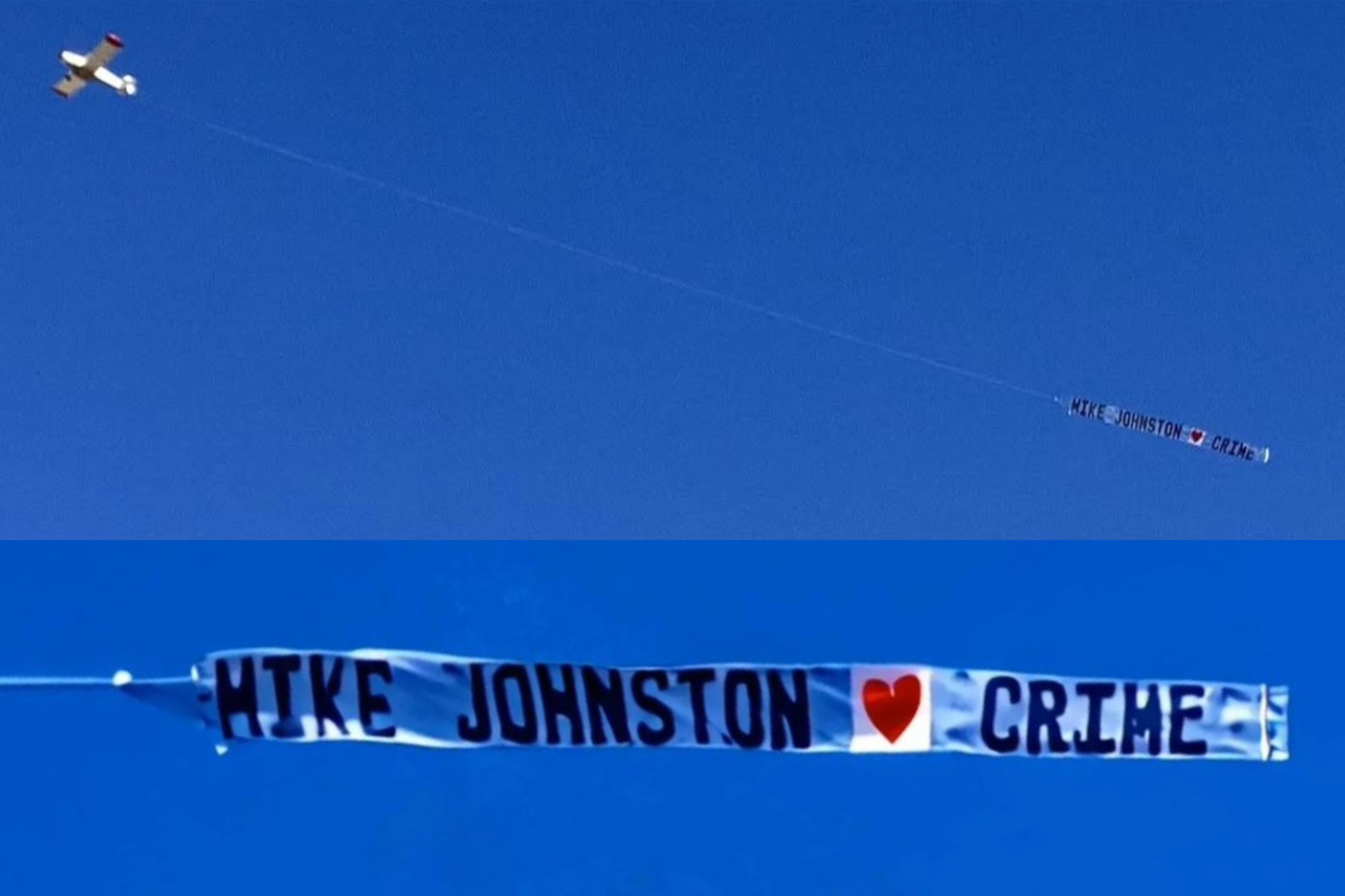 A small plane flies through the sky dragging a banner that reads "Mike Johnston [hearts] crime."
