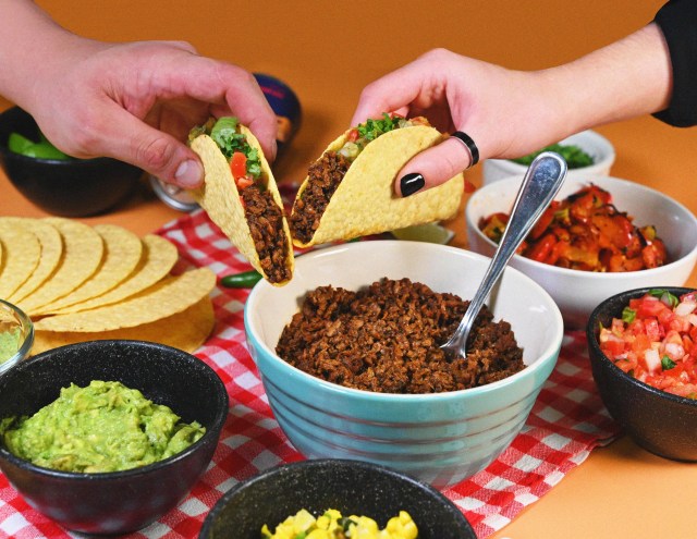 Two hands holding hard shell tacos over bowls of Illegal Pete's menu items