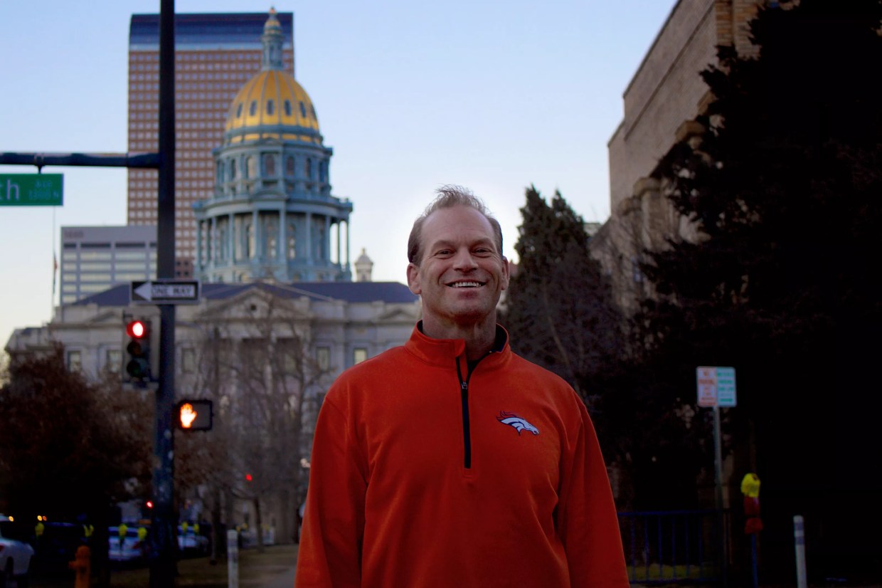 Jason Clark wearing an orange Broncos sweatshirt standing in front of the Colorado Capitol building.