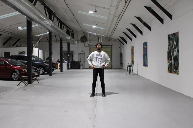 A Black man stands in the center of an art gallery/auto dealership.