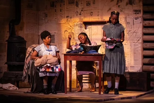 Two women sit, and one stands around a wooden table.