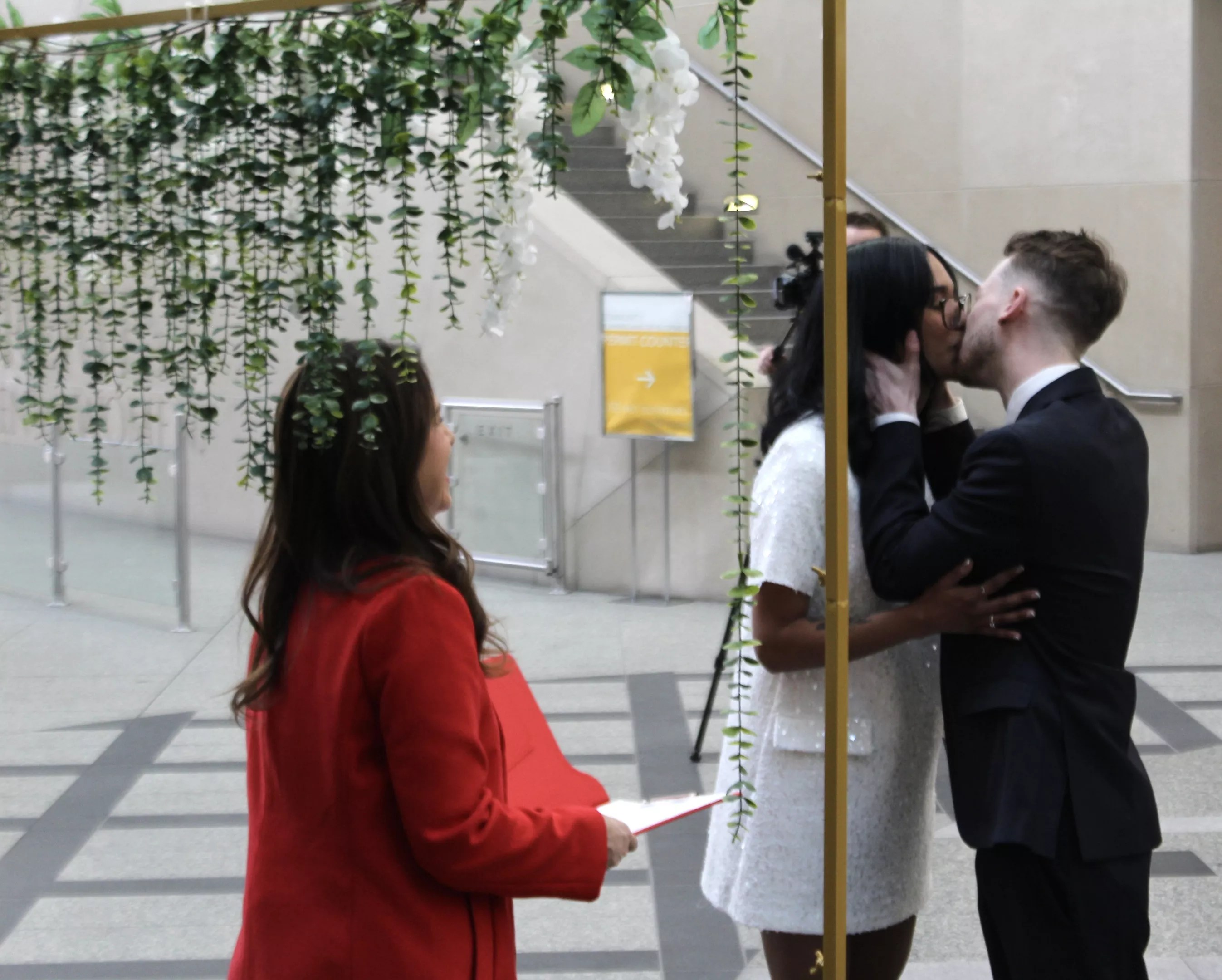 A man and woman kiss at the altar, an officiant wearing red looks on