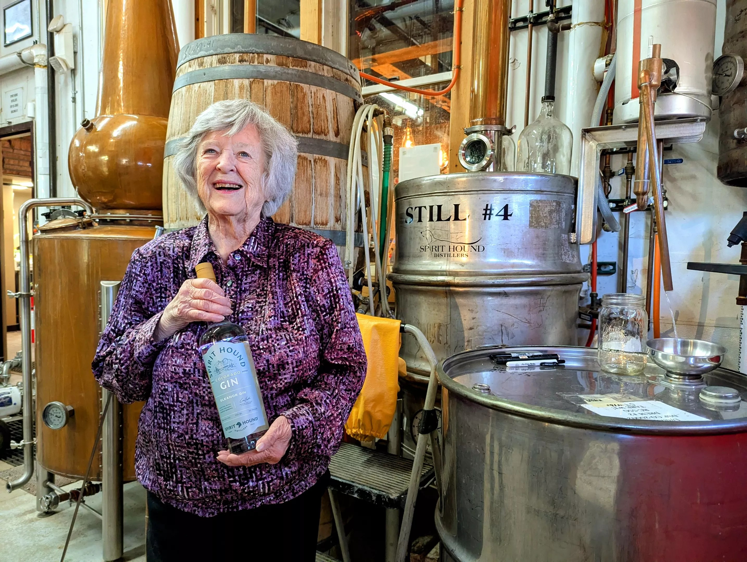 woman holding a bottle of Eleanor Gin while backdropped by equipment at Spirit Hound's distillery