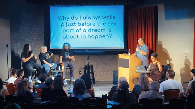 People on stage performing in front of a projection screen that reads, "Why do I always wake up just before the sex part of a dream is about to happen?"