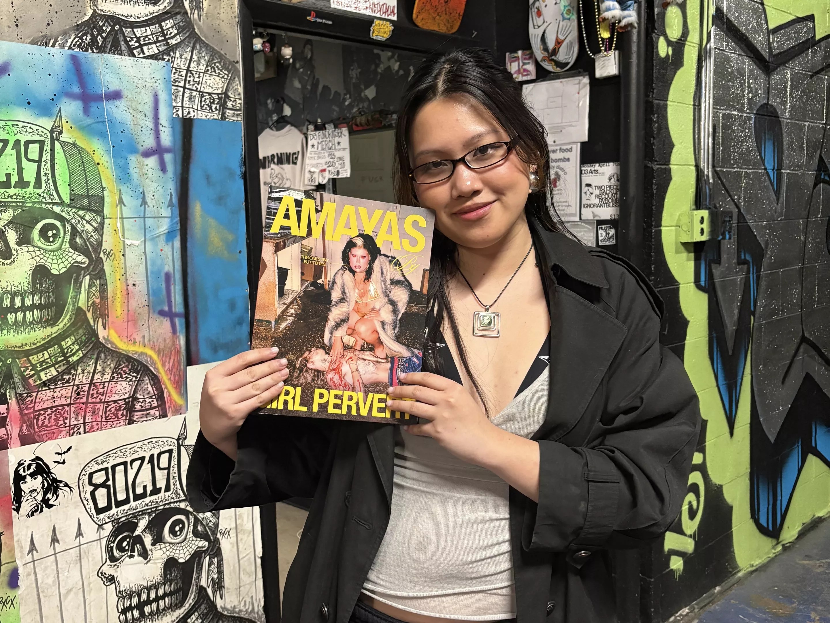 Hà Pham stands in front of a mural wall while holding a copy of Girl Pervert: Volume One with cover star Amayas Gonzalez.
